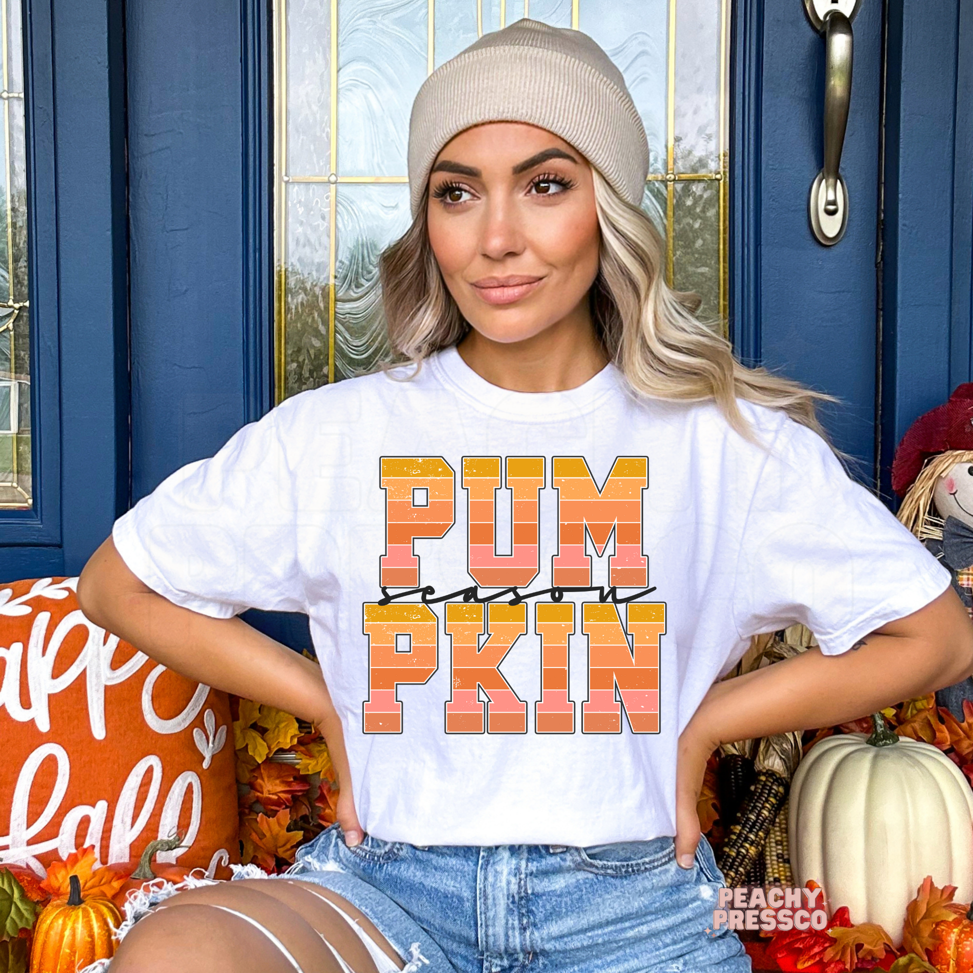 Woman wearing a 'PUMPKIN' shirt in front of a blue door with fall decorations.