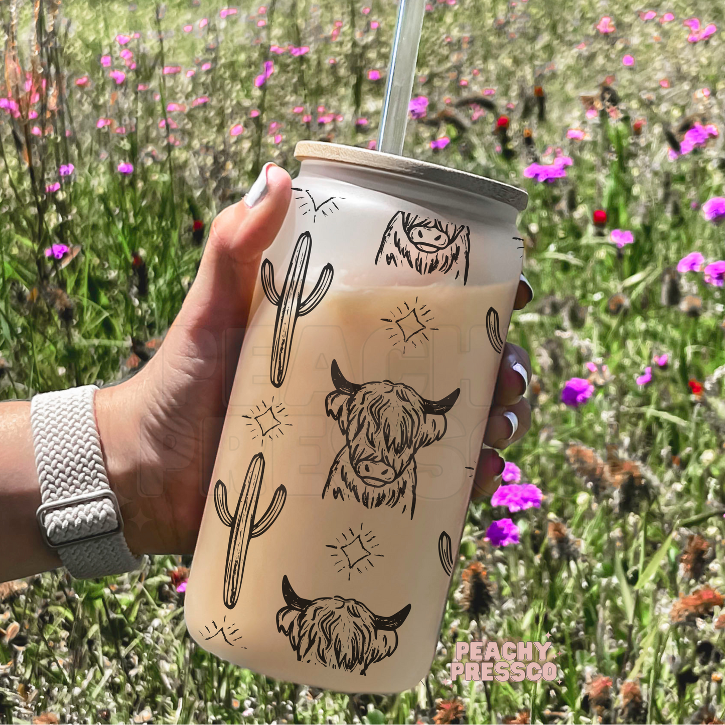 A hand holds a frosted tumbler with a straw in a grassy field with pink wildflowers. The tumbler has black illustrations of cacti, highland cows, and suns.