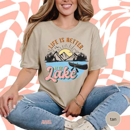 Woman sitting in ripped jeans wearing tan graphic t-shirt with colorful “Life Is Better at the Lake” outdoor design.
