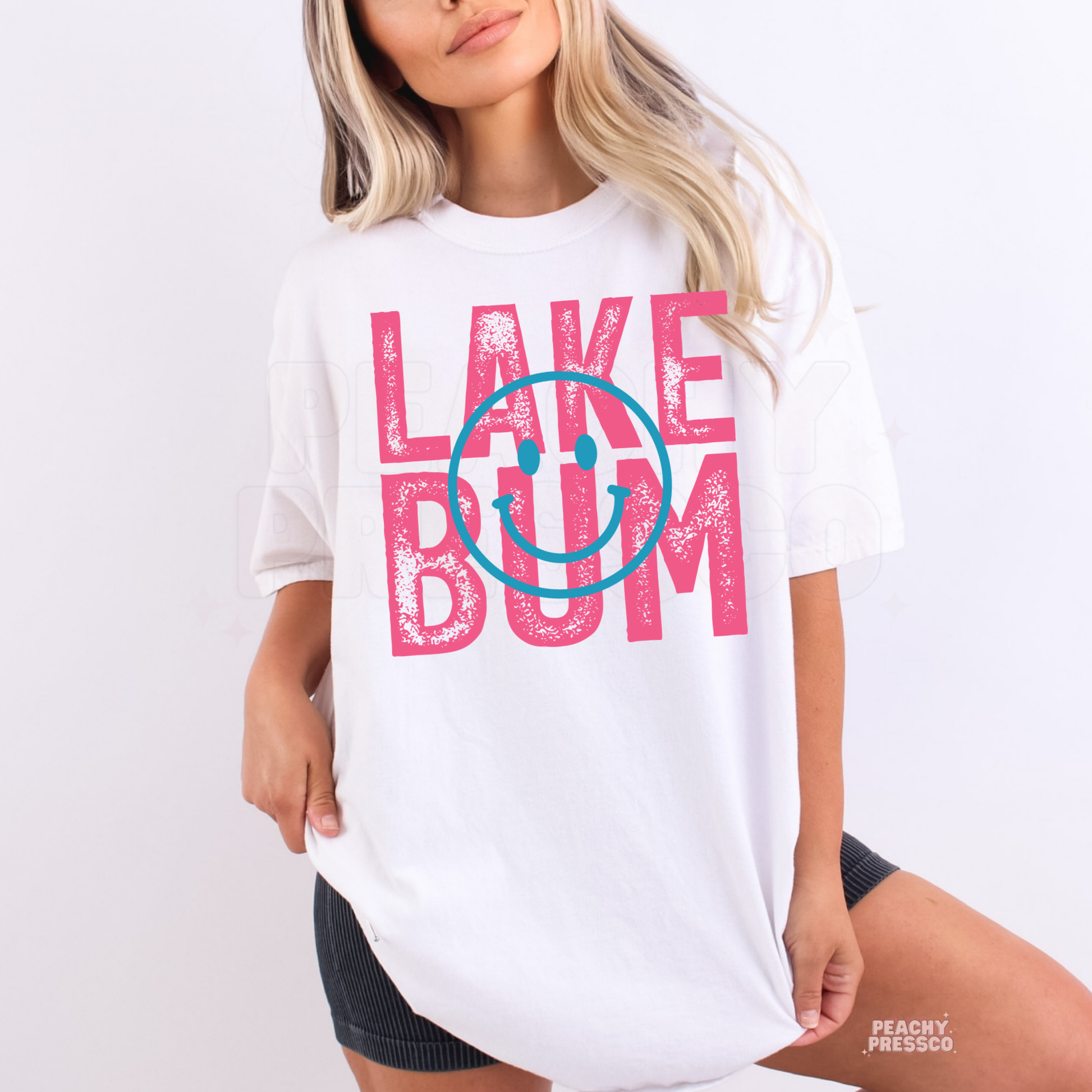 Woman wearing oversized white Lake Bum graphic t-shirt with pink retro lettering and blue smiley face design.