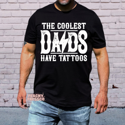 Man wearing black t-shirt with distressed “The Coolest Dads Have Tattoos” graphic in white western-style font.