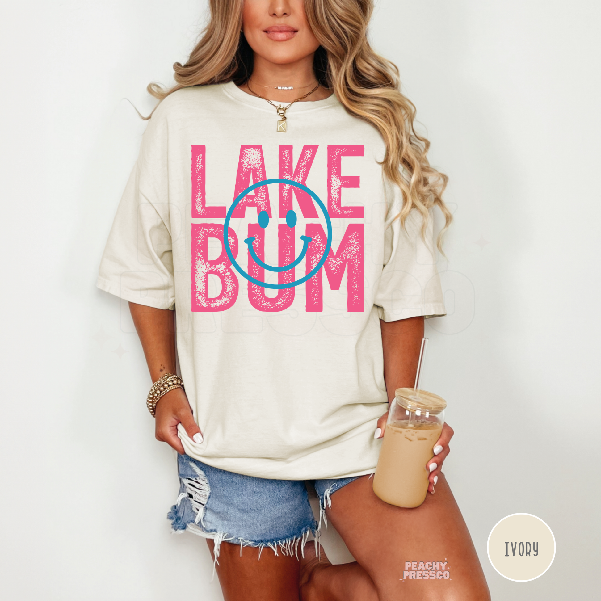 Woman wearing oversized ivory Lake Bum graphic t-shirt with pink retro lettering and blue smiley face while holding iced coffee.