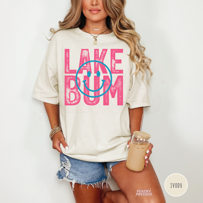 Woman wearing oversized ivory Lake Bum graphic t-shirt with pink retro lettering and blue smiley face while holding iced coffee.