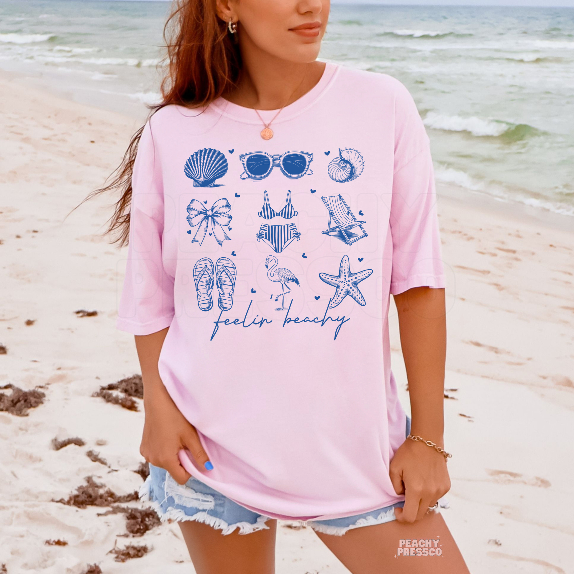 Woman wearing a light pink oversized beach t-shirt with blue coastal icons graphic featuring seashells, sunglasses, flip flops, flamingo, and “feelin beachy” text.