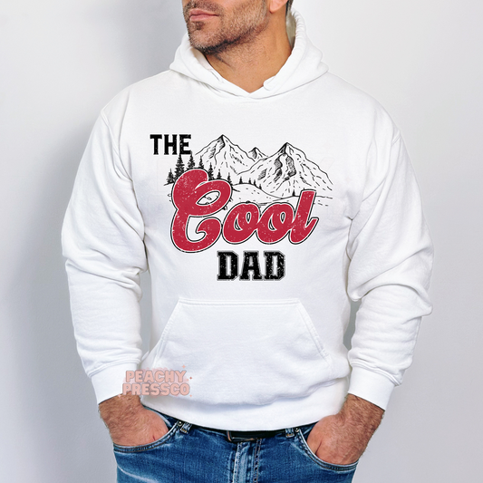 Man wearing white hoodie with “The Good Dad” graphic featuring red retro lettering and mountain landscape illustration.