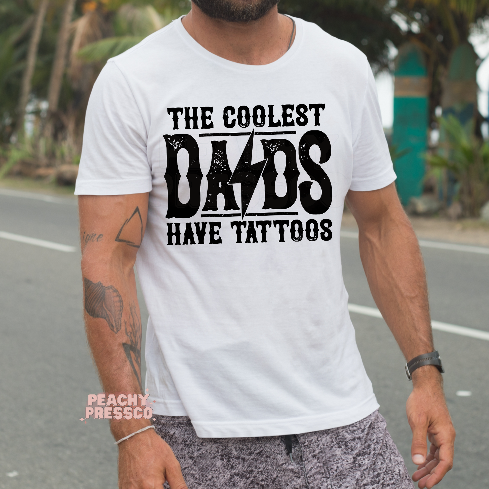 Man outdoors wearing white t-shirt with bold “The Coolest Dads Have Tattoos” graphic design.