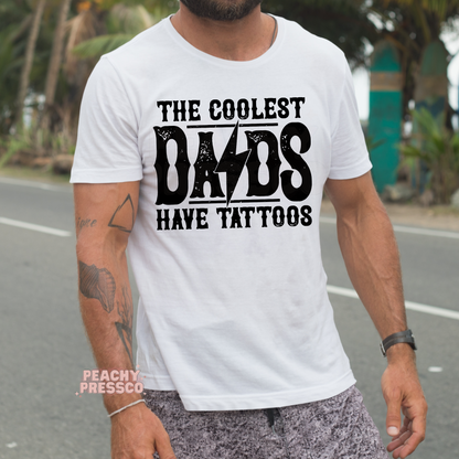 Man outdoors wearing white t-shirt with bold “The Coolest Dads Have Tattoos” graphic design.