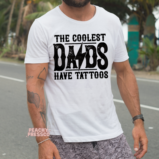 Man outdoors wearing white t-shirt with bold “The Coolest Dads Have Tattoos” graphic design.