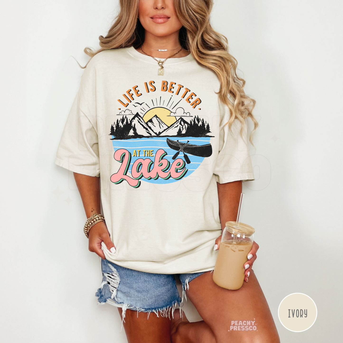 Woman wearing oversized ivory lake graphic t-shirt while holding iced coffee featuring canoe, mountain, and sunset artwork.