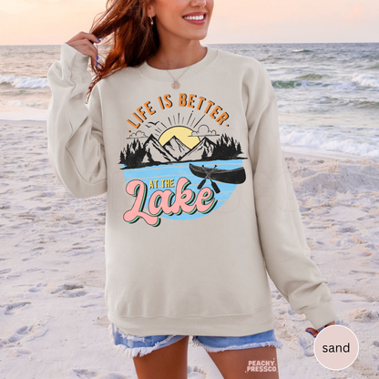 Woman wearing oversized sand colored sweatshirt on the beach with retro “Life Is Better at the Lake” graphic and canoe illustration.