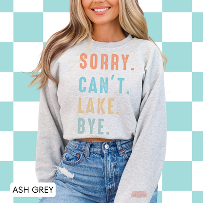 Woman wearing ash grey crewneck sweatshirt with colorful “Sorry. Can’t. Lake. Bye.” retro lake life graphic.
