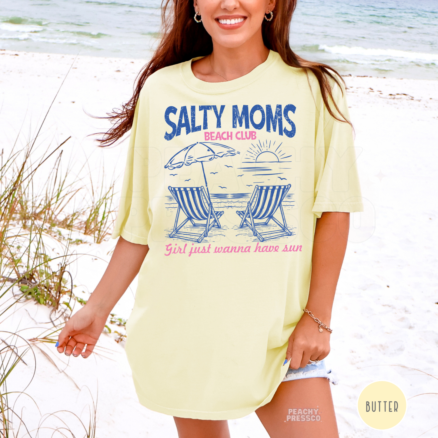 Woman wearing a butter yellow oversized beach t-shirt featuring a “Salty Moms Beach Club” graphic with striped beach chairs, umbrella, and sunset design.