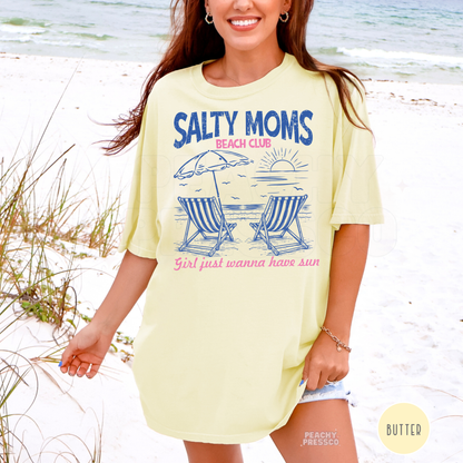 Woman wearing a butter yellow oversized beach t-shirt featuring a “Salty Moms Beach Club” graphic with striped beach chairs, umbrella, and sunset design.