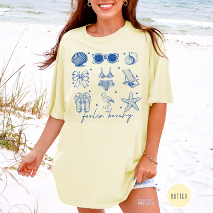 Woman wearing a butter yellow oversized beach t-shirt featuring blue coastal inspired icons and “feelin beachy” graphic print.