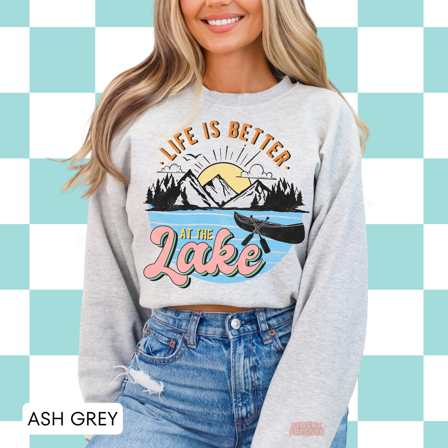 Woman wearing ash grey crewneck sweatshirt with retro “Life Is Better at the Lake” graphic featuring mountains and canoe.