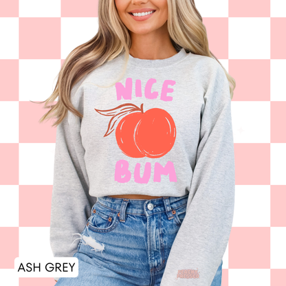 Woman wearing an ash grey sweatshirt featuring a peach illustration and pink “Nice Bum” graphic in a playful retro style.