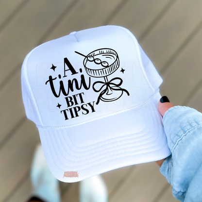 White trucker hat with black “A Tiny Bit Tipsy” cocktail graphic held outdoors, funny women’s hat for girls trips, bachelorette parties and summer outfits.
