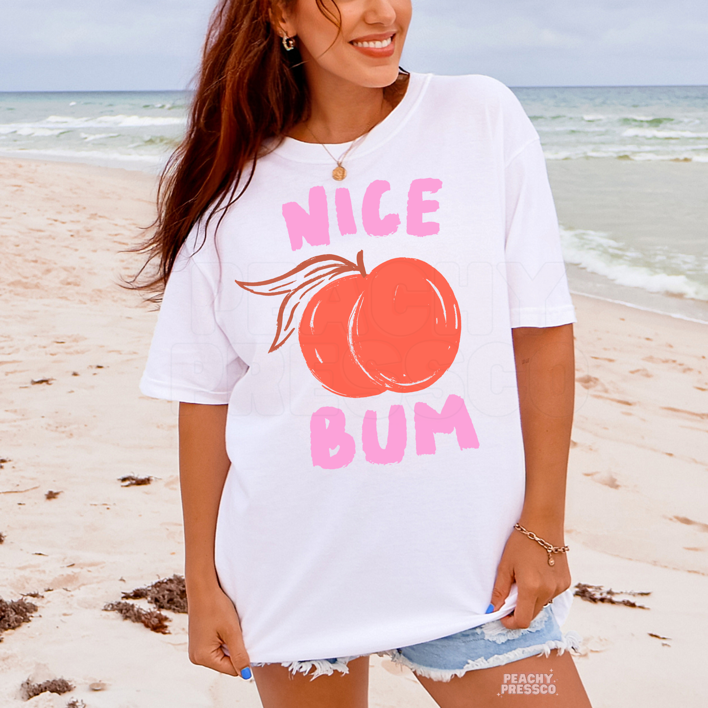 Woman wearing a white oversized beach t-shirt featuring a pink “Nice Bum” graphic with a peach illustration and playful retro lettering.