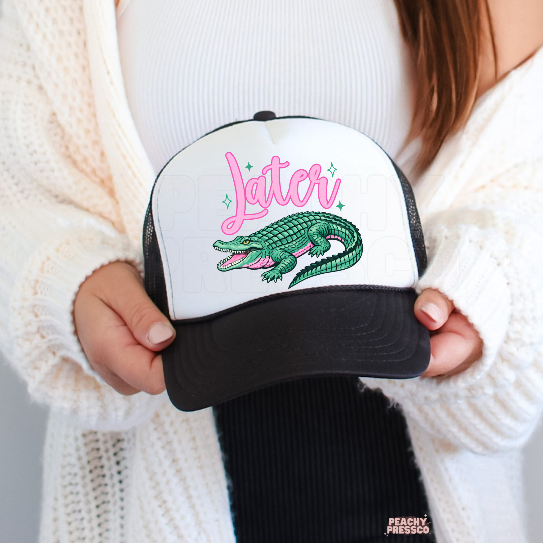 Woman holding a white and black trucker hat featuring a colorful alligator graphic and pink Later text.