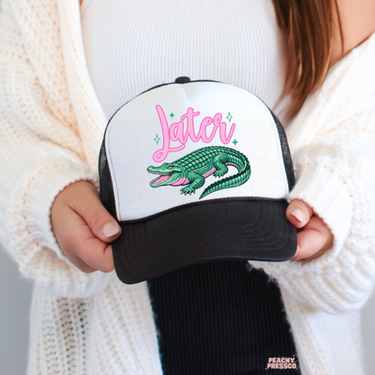 Woman holding a white and black trucker hat featuring a colorful alligator graphic and pink Later text.