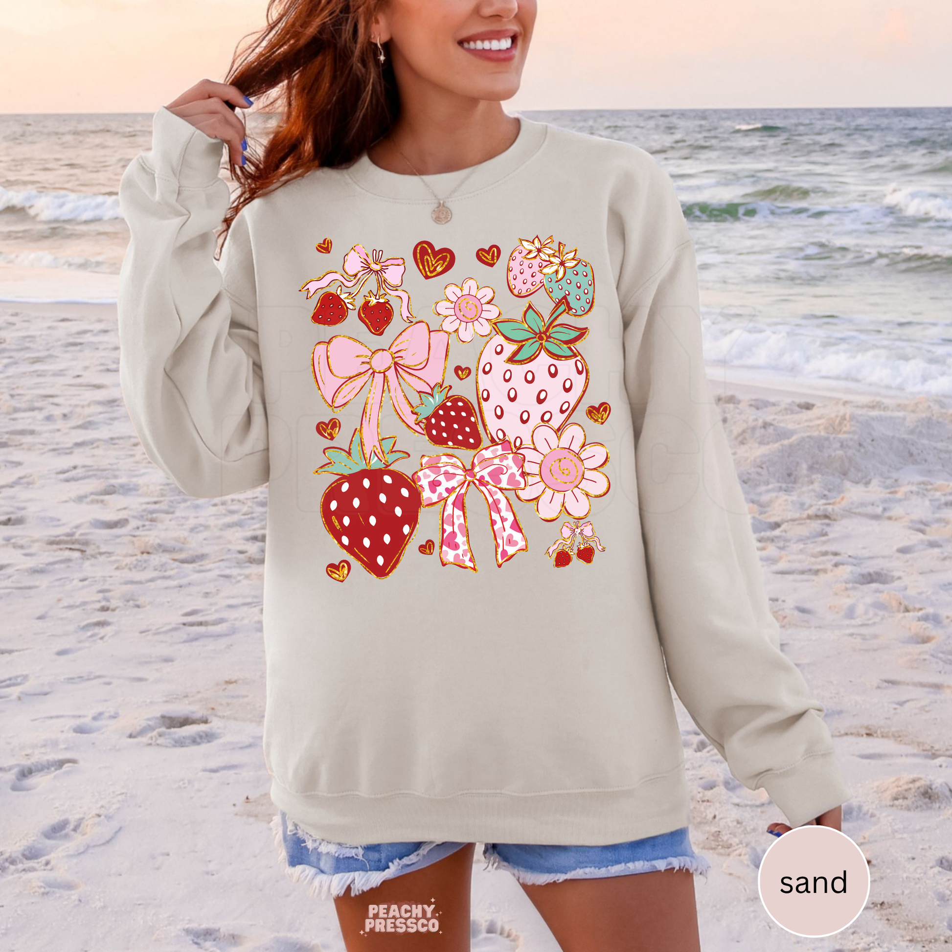 Woman wearing a sand-colored crewneck sweatshirt with a whimsical strawberry collage graphic featuring pink bows, flowers, and heart details.