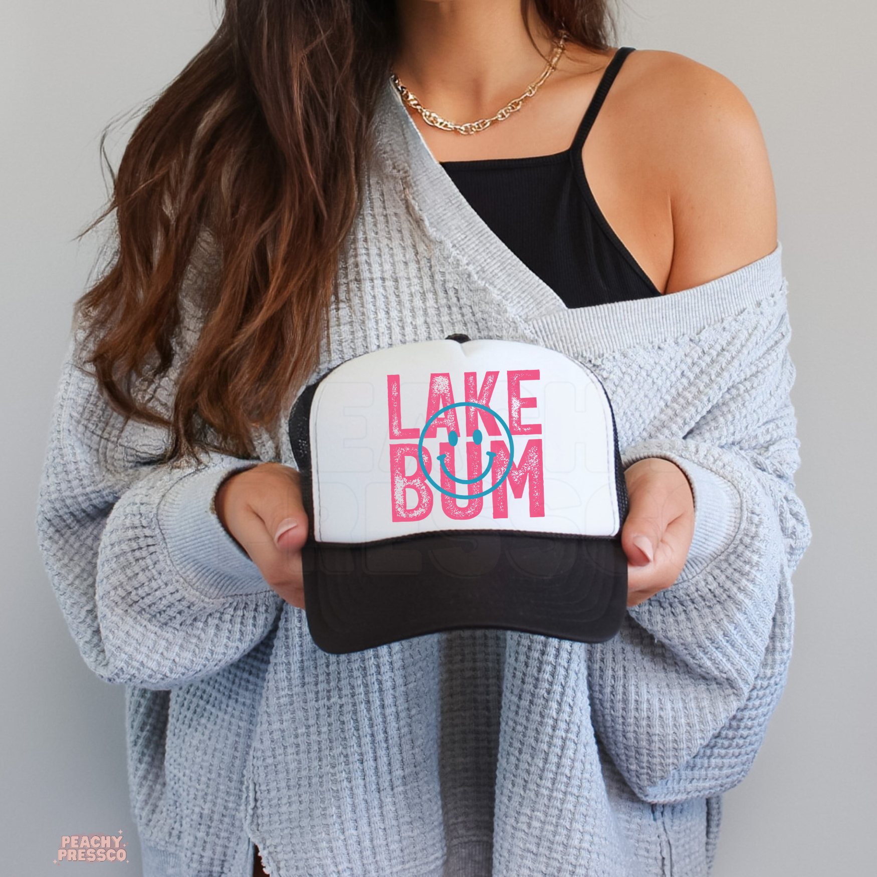 Woman holding a white and black trucker hat featuring pink Love Bum lettering with a retro smiley face design.