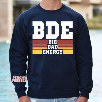 Man wearing navy crewneck sweatshirt featuring bold “BDE Big Dad Energy” retro dad graphic design.