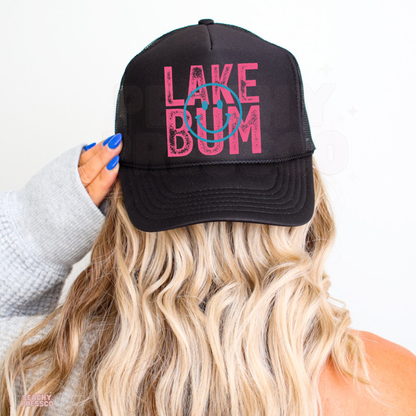 Black trucker hat with distressed pink “Lake Bum” text and turquoise smiley face graphic, styled for summer, beach and lake life outfits.