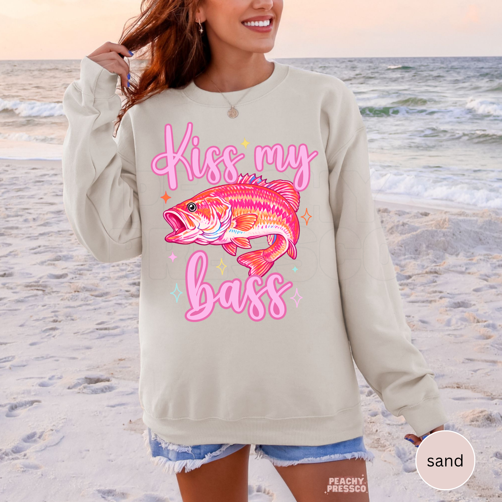 Woman wearing a sand crewneck sweatshirt featuring a colorful pink bass fish graphic with “Kiss My Bass” text, styled for lake days, fishing trips and summer outfits.