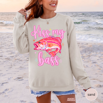 Woman wearing a sand crewneck sweatshirt featuring a colorful pink bass fish graphic with “Kiss My Bass” text, styled for lake days, fishing trips and summer outfits.