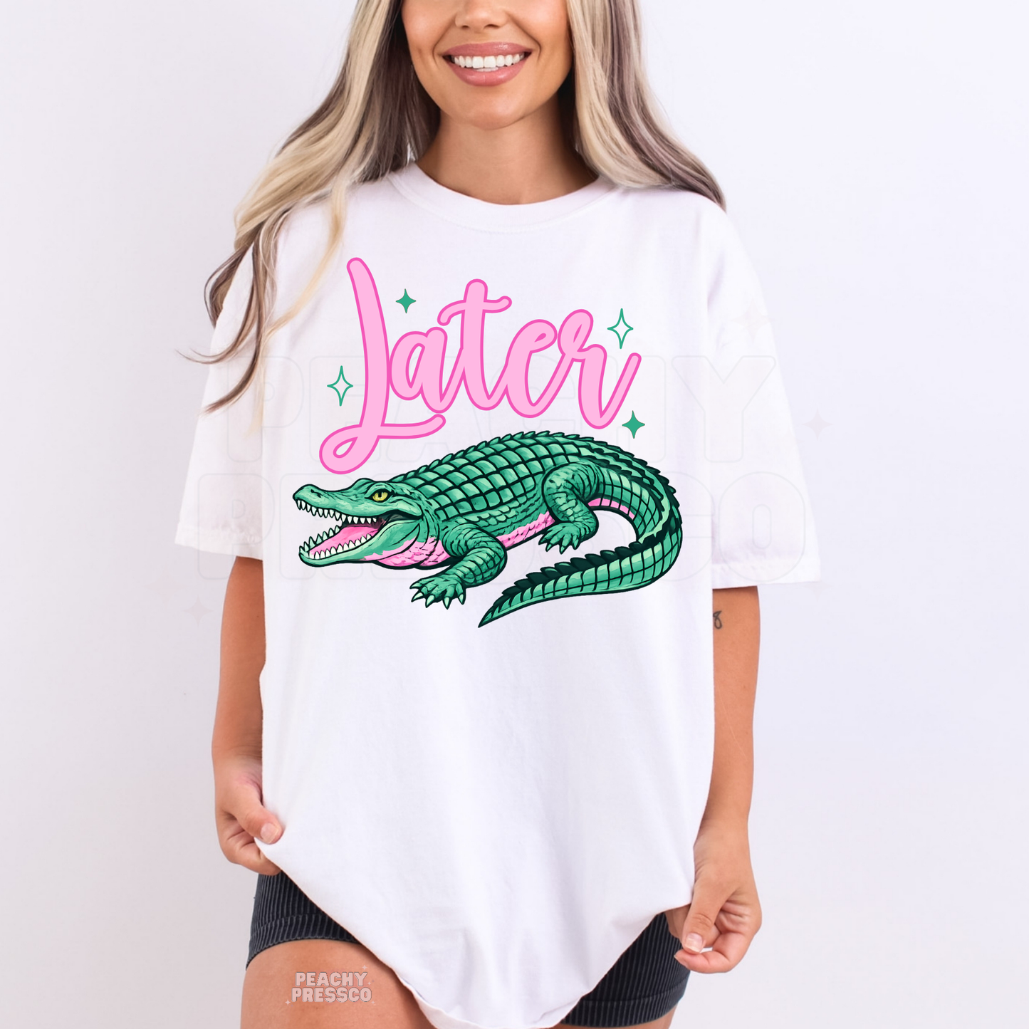 Woman wearing a white graphic t-shirt featuring a colorful alligator illustration with pink “Later” lettering and sparkles.
