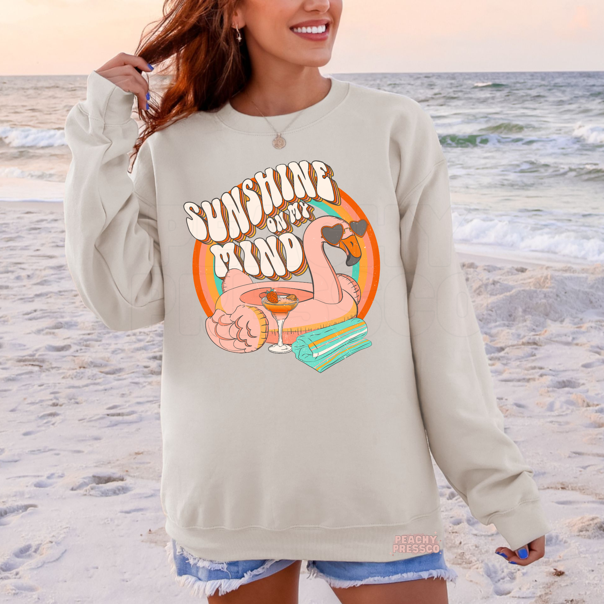 Woman wearing a sand colored crewneck sweatshirt with colorful flamingo float and rainbow graphic reading “Sunshine on My Mind.”