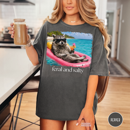 Woman wearing a pepper oversized graphic t-shirt featuring a sunglasses raccoon relaxing on a pink float with tropical ocean artwork and “Feral and Salty” design.