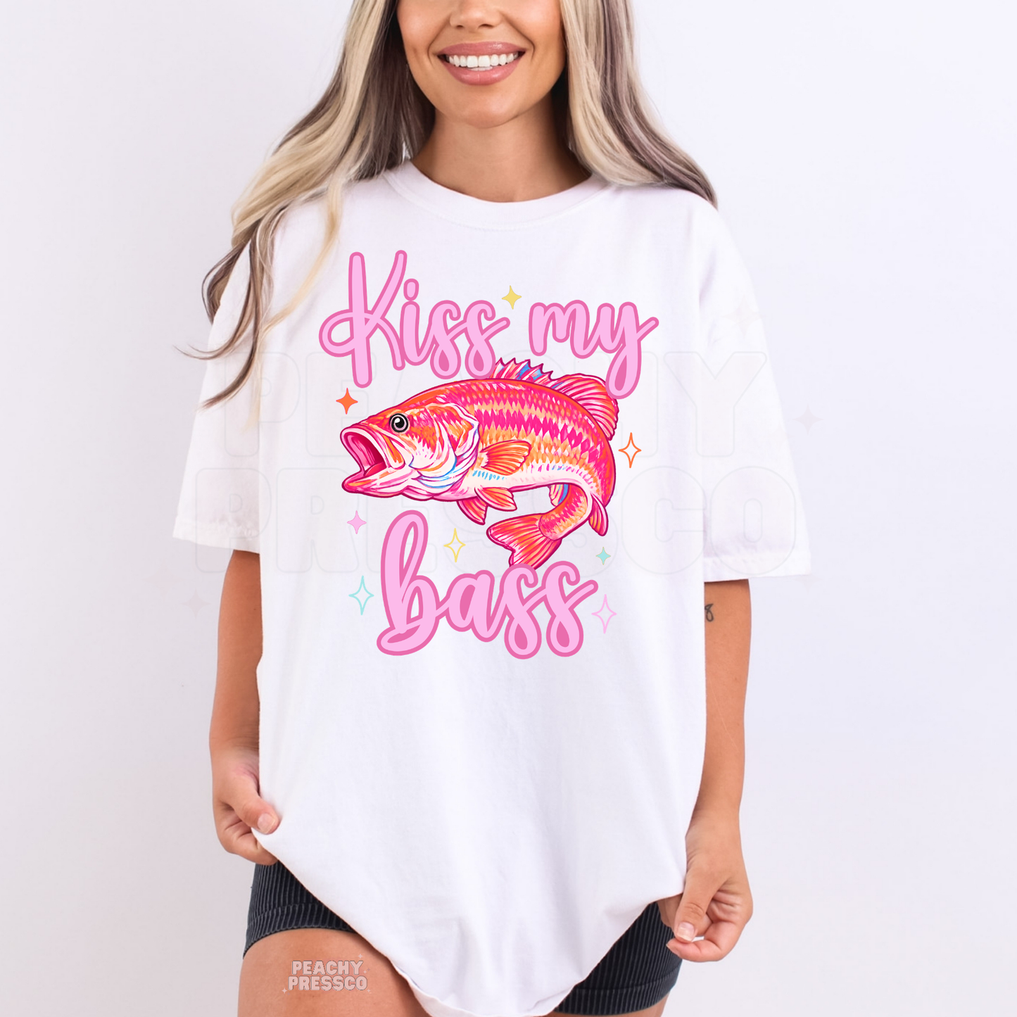 Woman wearing a white graphic t-shirt featuring colorful bass fish artwork with pink “Kiss My Bass” lettering.
