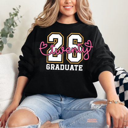 Woman wearing a black Twenty 26 Graduate sweatshirt featuring white and gold varsity numbers, pink script lettering, and Class of 2026 graduation design.