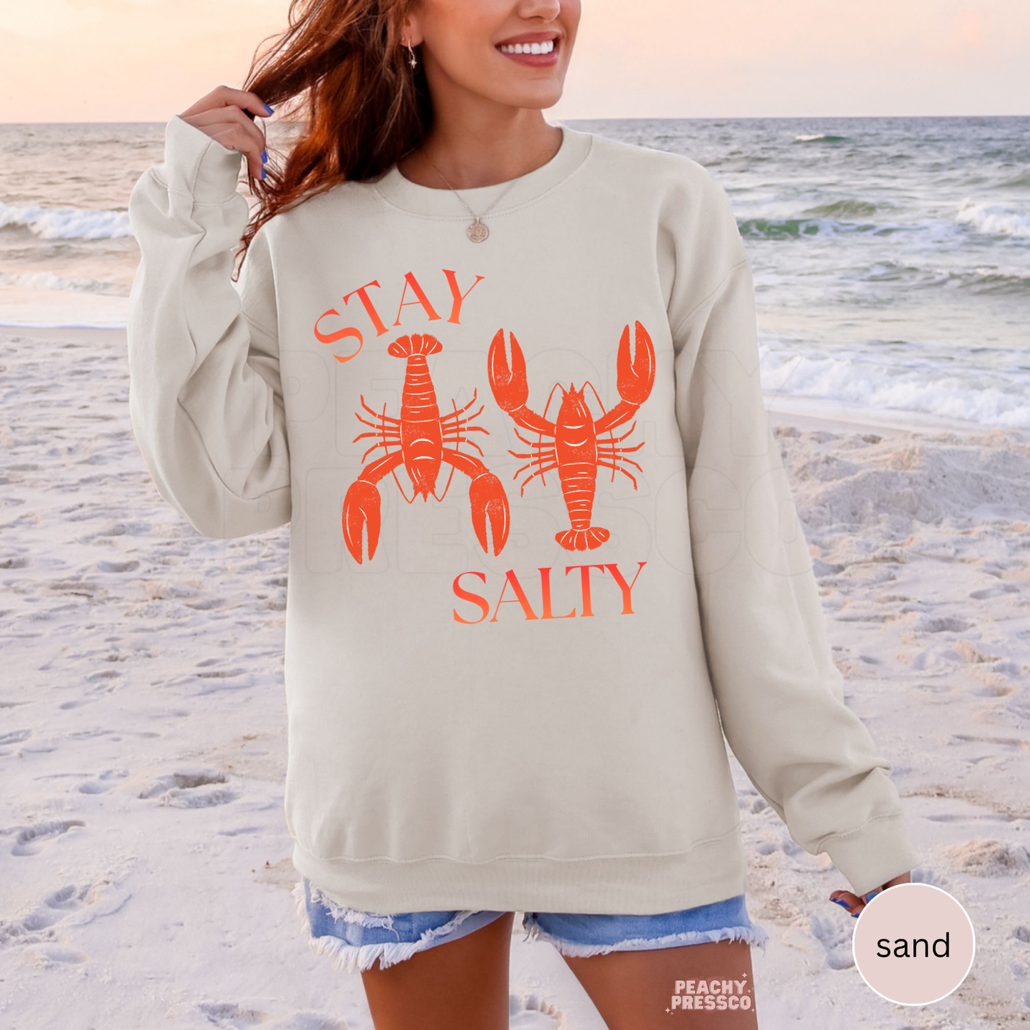 Sand crewneck sweatshirt featuring coral “Stay Salty” lobster graphic – coastal beach sweatshirt for women, summer vacation apparel.
