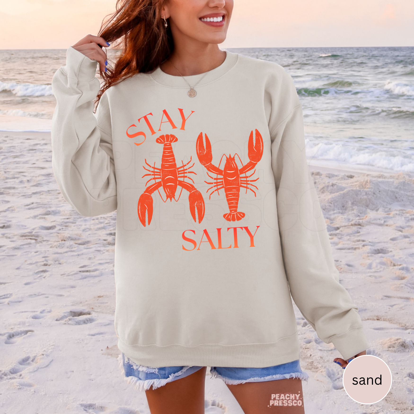 Sand crewneck sweatshirt featuring coral “Stay Salty” lobster graphic – coastal beach sweatshirt for women, summer vacation apparel.