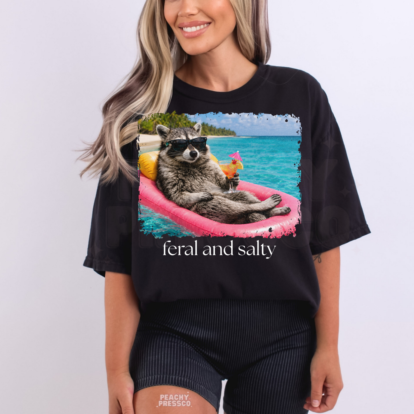 Woman wearing a black graphic t-shirt featuring a raccoon floating on a pink raft with tropical drink and “Feral and Salty” beach graphic.