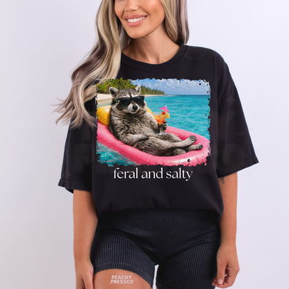 Woman wearing a black graphic t-shirt featuring a raccoon floating on a pink raft with tropical drink and “Feral and Salty” beach graphic.