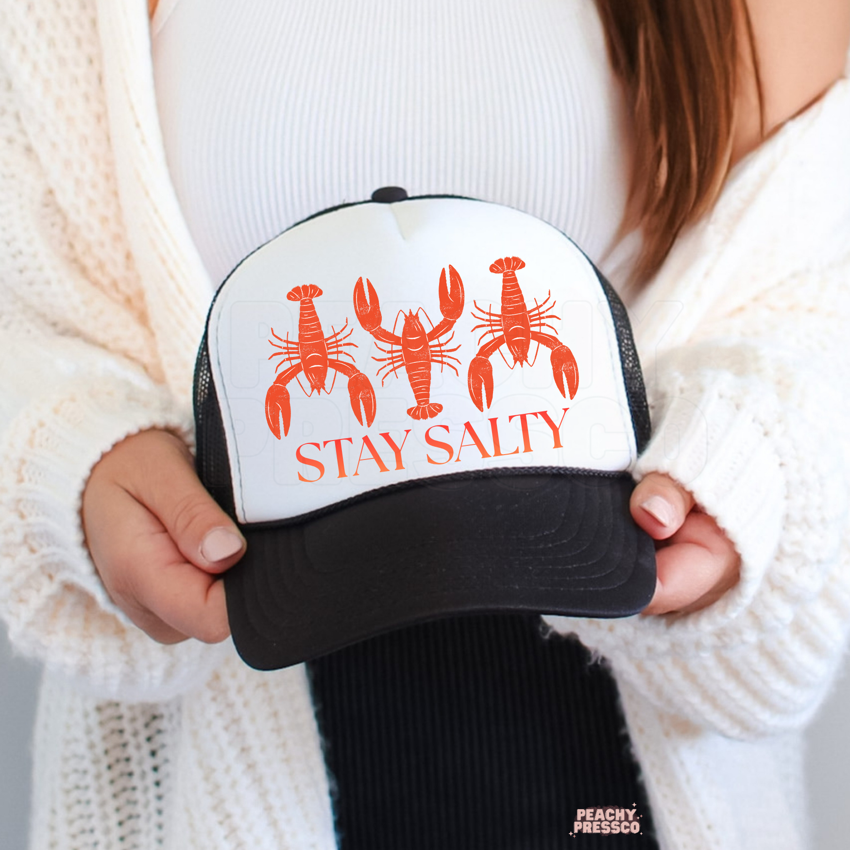 Woman holding a white and black trucker hat featuring orange lobster illustrations and the text Stay Salty.