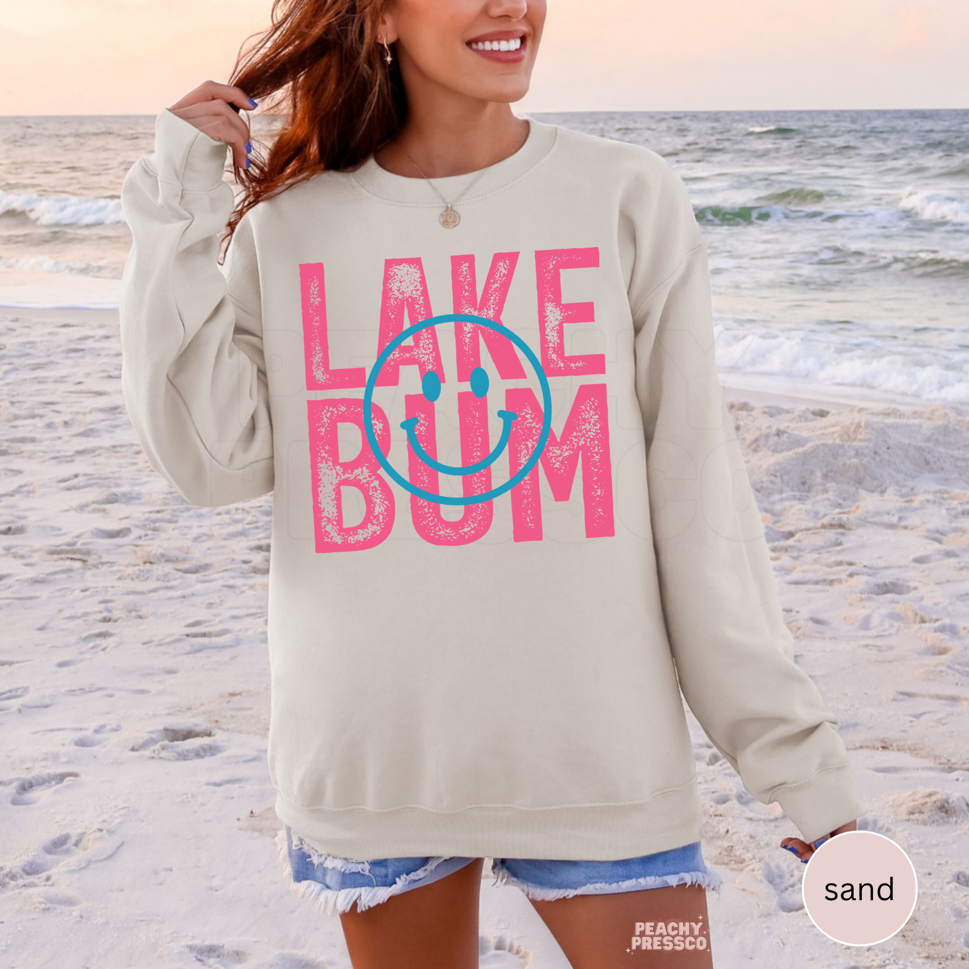 Woman wearing oversized sand colored Lake Bum sweatshirt at the beach with pink retro text and blue smiley face graphic.