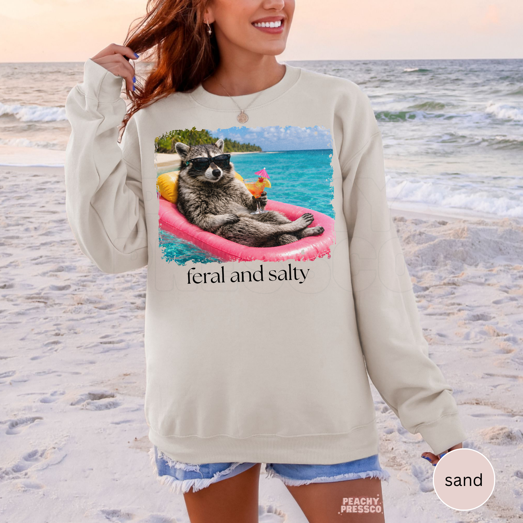 Woman wearing a sand colored crewneck sweatshirt featuring a funny raccoon float graphic with tropical beach scene and “Feral and Salty” text.