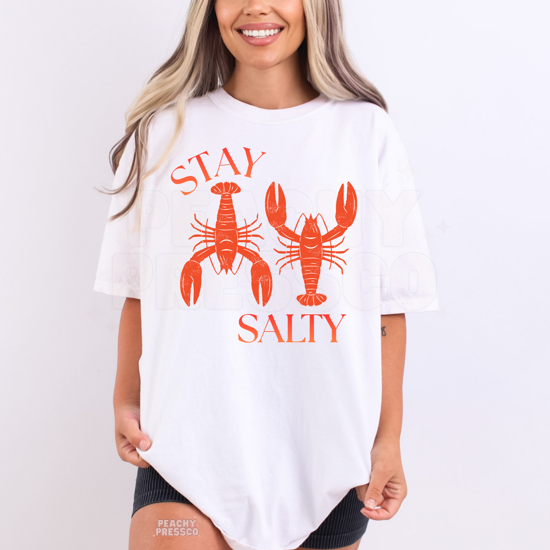Woman wearing a white graphic t-shirt featuring coral lobster illustrations with “Stay Salty” text and coastal summer style.
