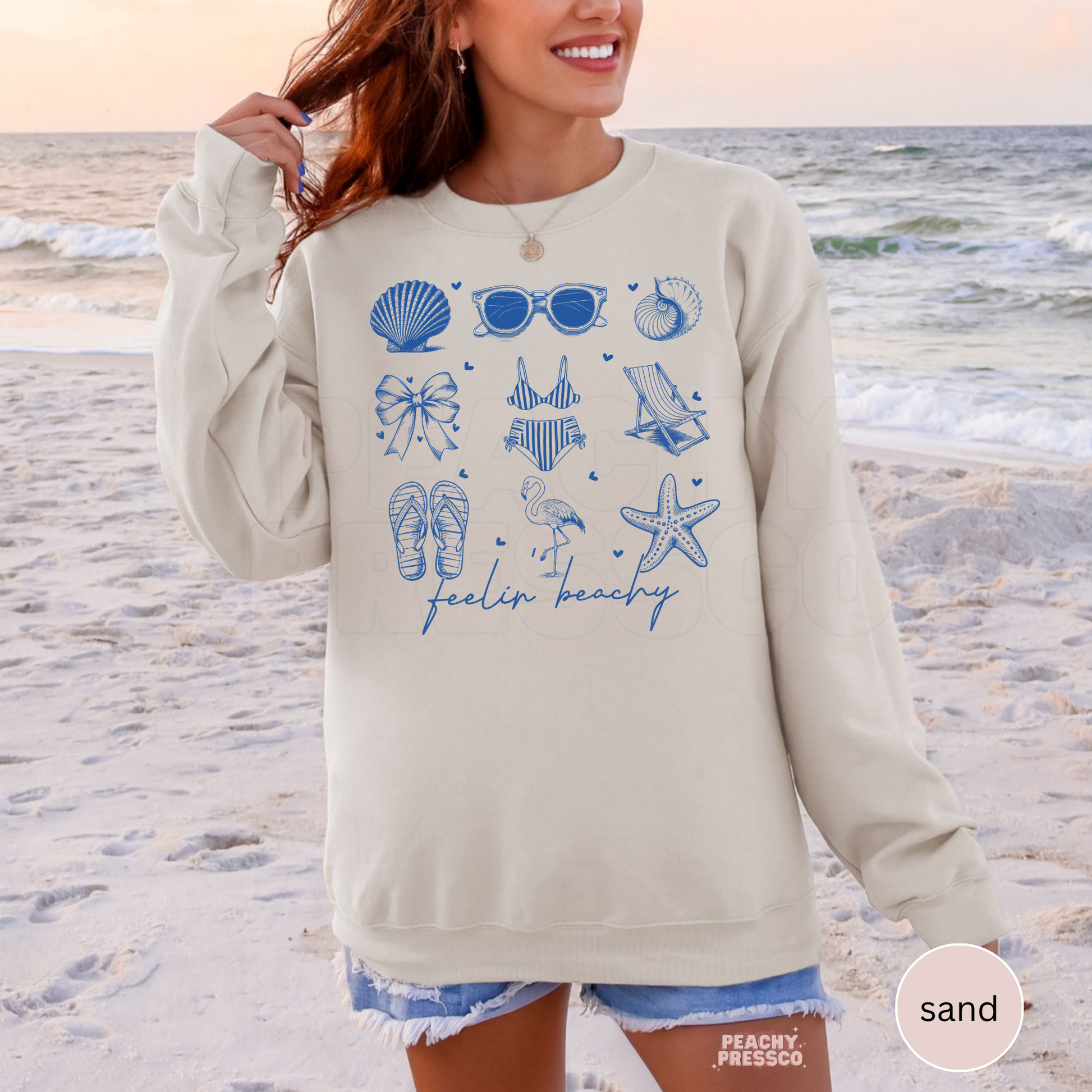 Woman wearing a sand colored beach sweatshirt with blue seaside doodle graphics and “feelin beachy” script design.