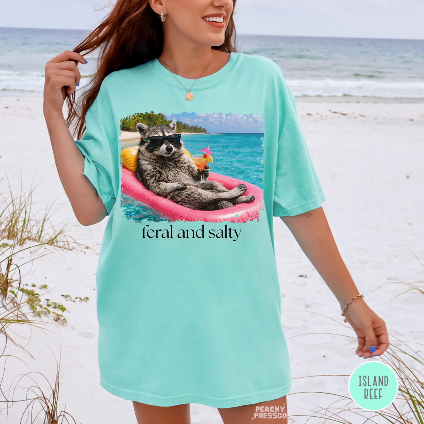 Woman wearing an island reef oversized graphic t-shirt featuring a raccoon floating on a pink raft holding a tropical drink with “Feral and Salty” text at the beach.
