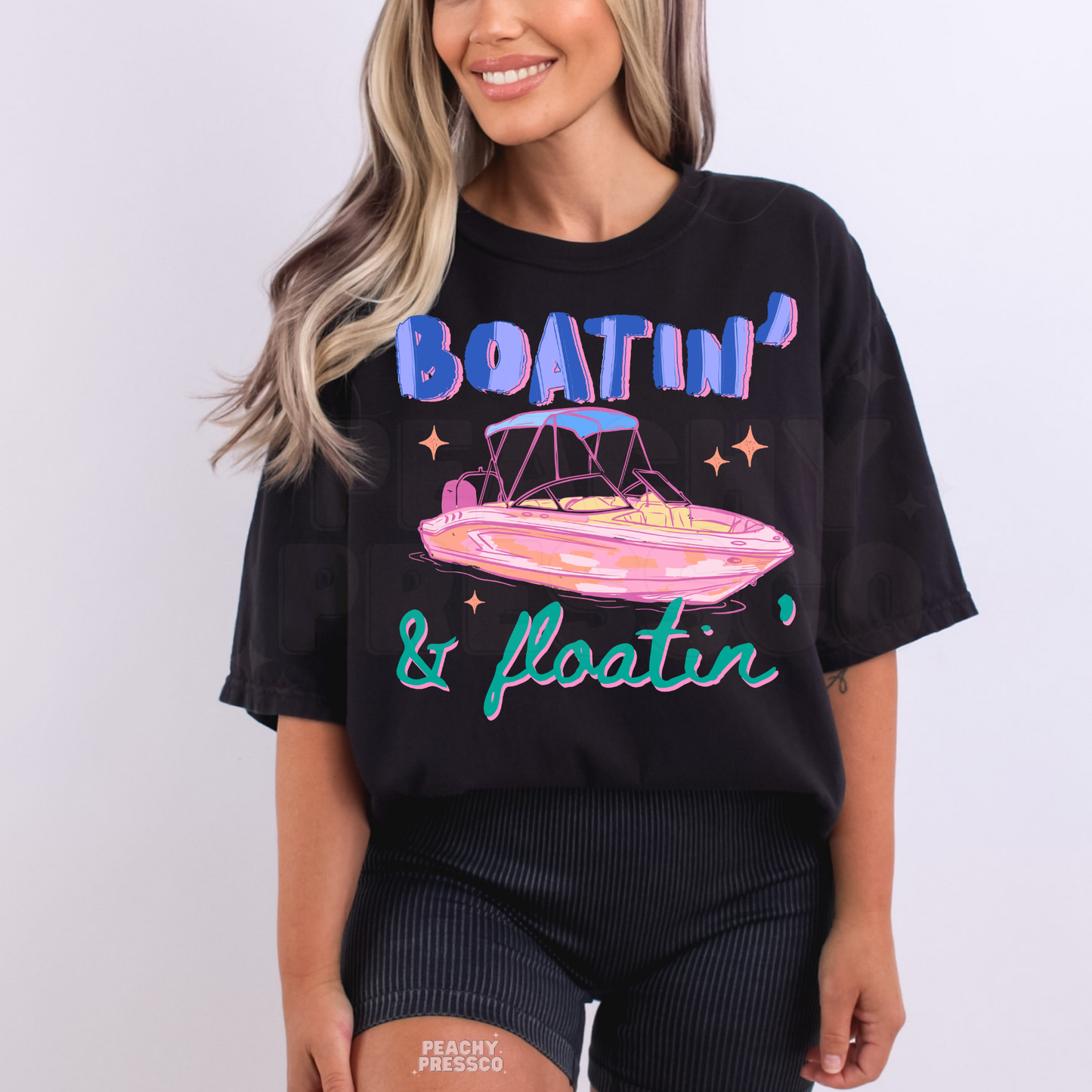 Woman wearing a black graphic t-shirt featuring colorful “Boatin’ & Floatin’” lettering with retro pink boat artwork and summer lake vibes.