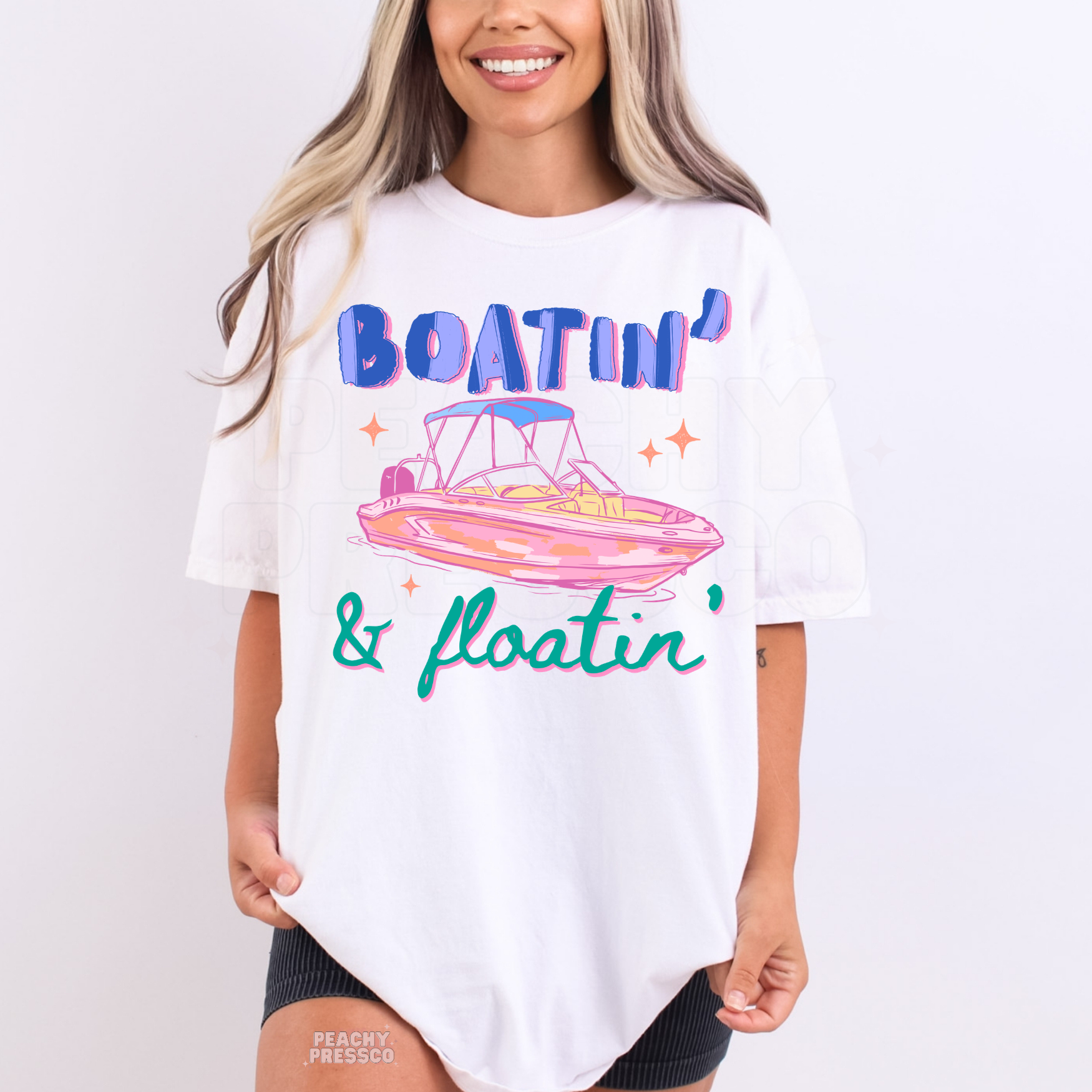 Woman wearing a white graphic t-shirt featuring colorful “Boatin’ & Floatin’” text with retro boat artwork inspired by summer lake life.