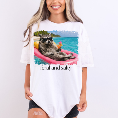 Woman wearing a white graphic t-shirt featuring a raccoon floating on a pink raft with tropical drink and “Feral and Salty” text.