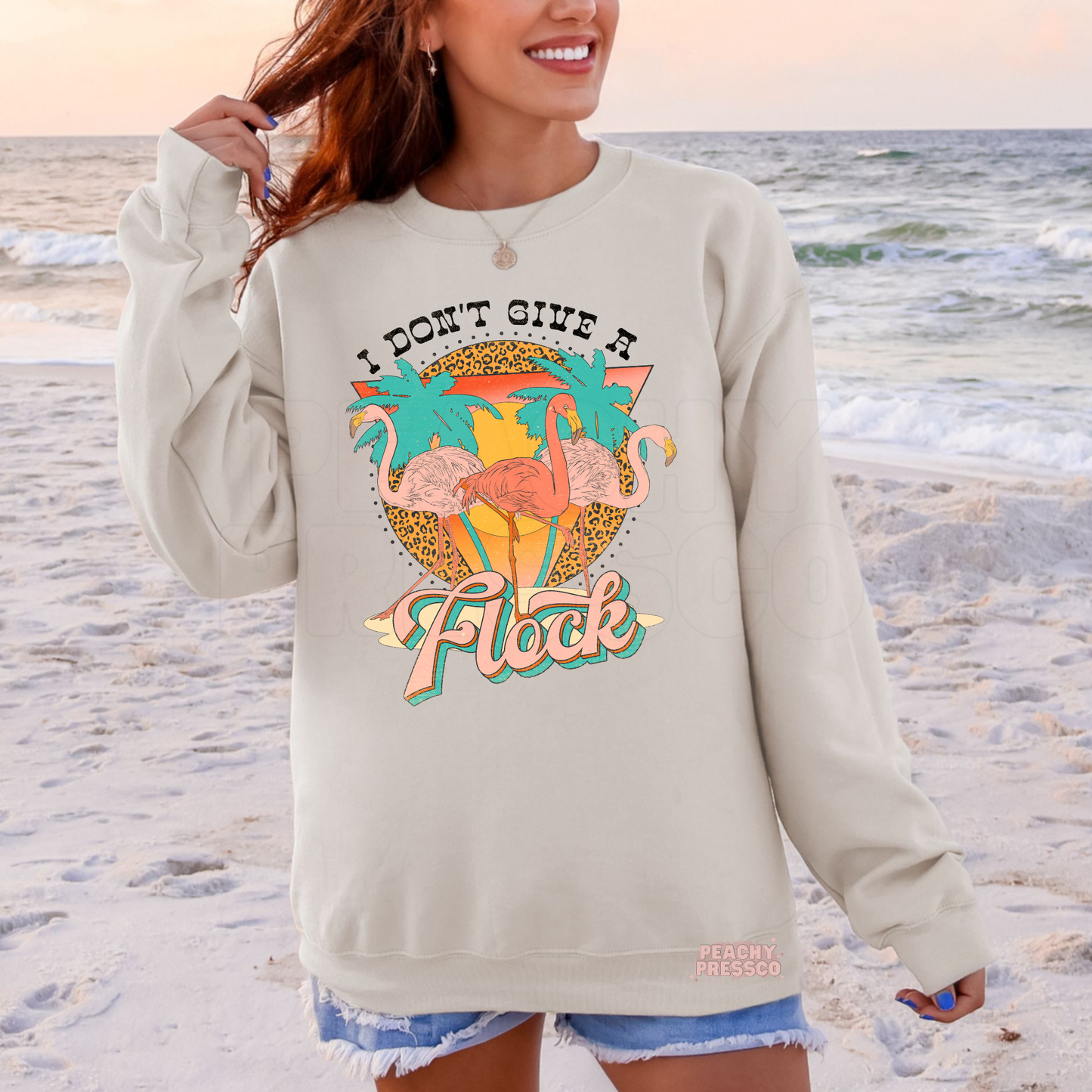 Woman wearing a sand colored beach sweatshirt featuring retro flamingos, palm trees, and “I Don’t Give A Flock” graphic.
