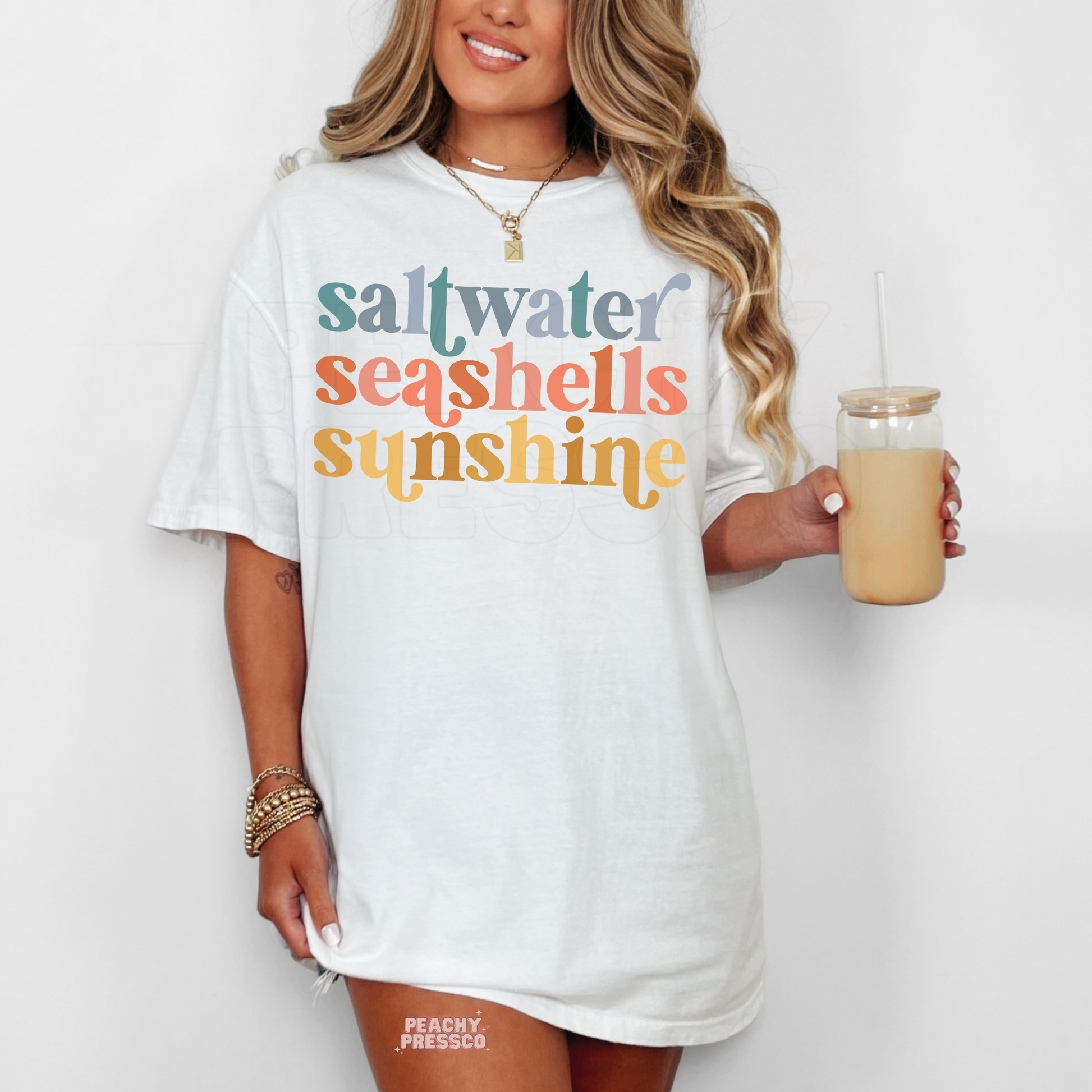 Woman wearing a white oversized t-shirt dress with colorful retro “saltwater seashells sunshine” typography and a beachy summer aesthetic.